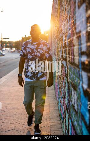 A Black man walks past a colorful mural in an urban setting as the sun goes down behind him. Stock Photo