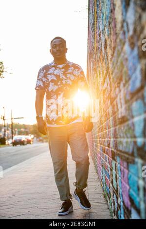 A Black man walks past a colorful mural in an urban setting as the sun goes down behind him. Stock Photo