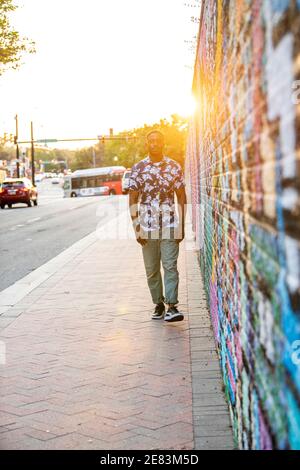A Black man walks past a colorful mural in an urban setting as the sun goes down behind him. Stock Photo