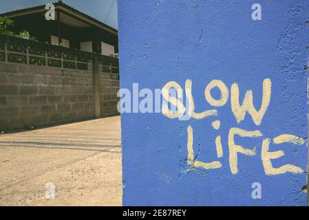 Blue painted wall with slow life written in white paint against view of old brick wall and empty street Stock Photo