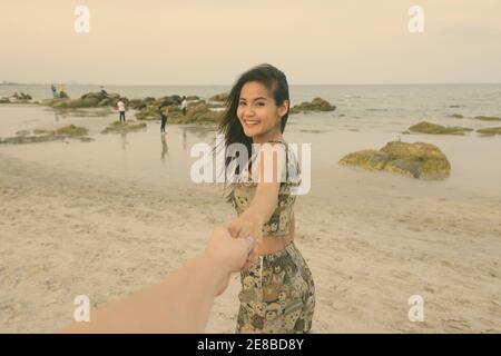 Young happy beautiful Asian woman smiling while holding hands and leading man at the public beach of Hua Hin in Thailand Stock Photo