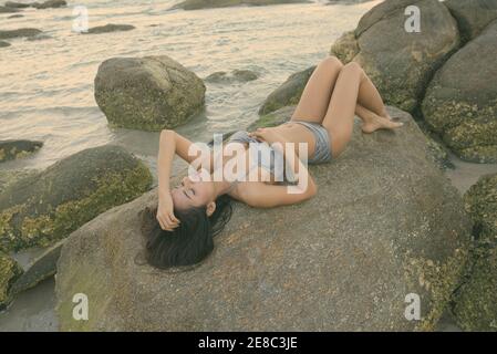 Young beautiful Asian woman lying down while posing with eyes closed on the rocks of public beach in Hua Hin Thailand Stock Photo