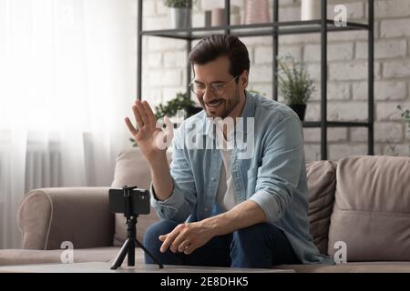 Smiling man wearing glasses waving hand, recording vlog at home Stock Photo