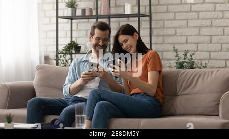 Overjoyed couple using phones together, sitting on cozy couch Stock Photo