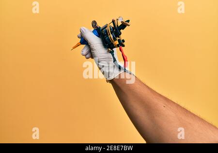 Hand of young hispanic man using tattoo machine over isolated yellow background. Stock Photo