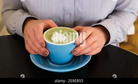 Woman holding blue cup with hot trendy green latte with art heart on the foam on black wooden table background, healthy drink with latte art, morning Stock Photo