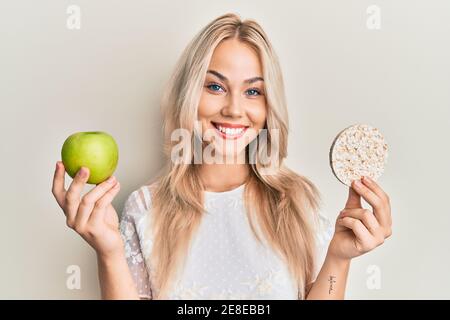 Beautiful caucasian blonde girl holding nachos and healthy green apple smiling with a happy and cool smile on face. showing teeth. Stock Photo