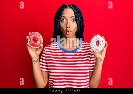 Beautiful hispanic woman holding tasty colorful doughnuts making fish face with mouth and squinting eyes, crazy and comical. Stock Photo