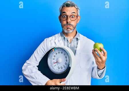Middle age grey-haired man as nutritionist doctor holding weighing machine and green apple puffing cheeks with funny face. mouth inflated with air, ca Stock Photo