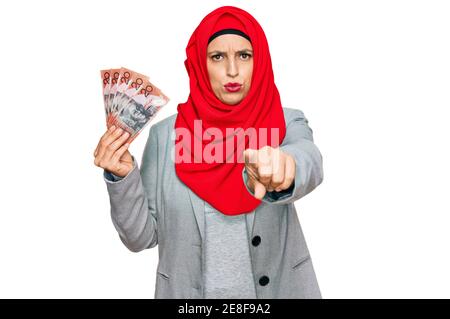 Beautiful hispanic woman wearing islamic hijab holding australian dollars banknotes pointing with finger to the camera and to you, confident gesture l Stock Photo