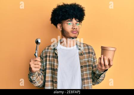 Young african american man with afro hair holding ice cream and ice cream scoop clueless and confused expression. doubt concept. Stock Photo