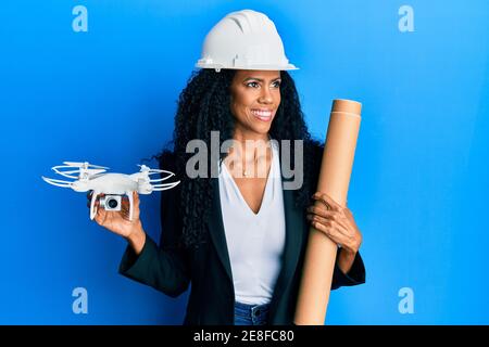 Middle age african american woman wearing safety helmet holding blueprints and drone smiling looking to the side and staring away thinking. Stock Photo