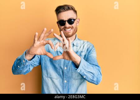 Young redhead man wearing stylish sunglasses smiling in love showing heart symbol and shape with hands. romantic concept. Stock Photo