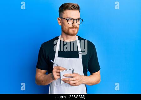 Young redhead man wearing waiter apron taking order smiling looking to the side and staring away thinking. Stock Photo