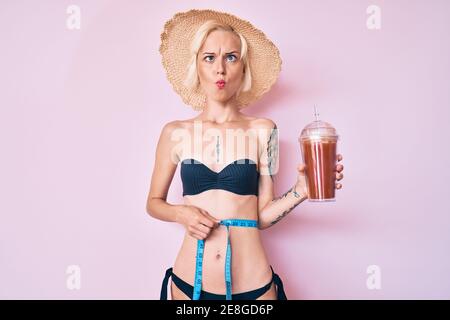 Young blonde woman with tattoo wearing bikini using tape measure and drinking smoothie making fish face with mouth and squinting eyes, crazy and comic Stock Photo