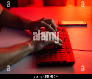 Crop anonymous male typing on keyboard while working on computer and sitting at desk with smartphone in dark room Stock Photo