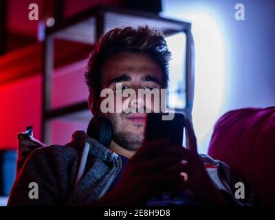 Content young male in casual wear and wireless headphones browsing mobile phone while resting on sofa in dark room Stock Photo