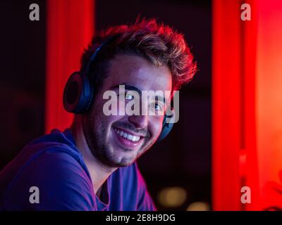 handsome happy male wearing casual shirt and wireless headphones leaning on desk with computer and looking at camera with smile in dark room Stock Photo