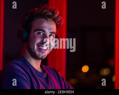 handsome happy male wearing casual shirt and wireless headphones leaning on desk with computer and looking at camera with smile in dark room Stock Photo