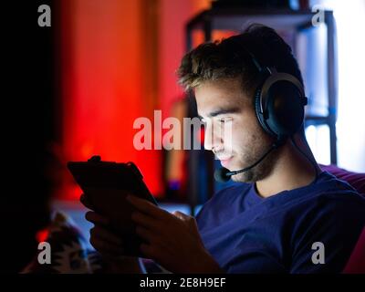 Content young male in casual wear and wireless headphones playing video games on tablet while resting on sofa in dark room Stock Photo