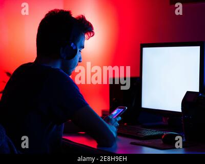 Side view focused young male in casual shirt and wireless headphones browsing mobile phone and working on computer with empty screen while sitting at Stock Photo