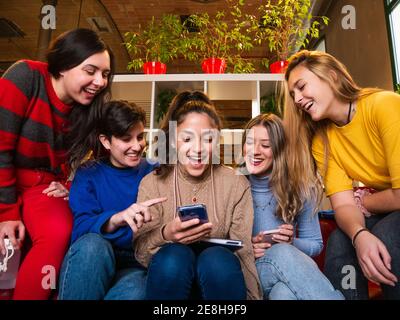 Smiling female students sitting on sofa with smartphone and having fun during break Stock Photo