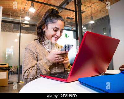 Low angle of smiling female student text messaging on cellphone while using laptop for studies in university Stock Photo