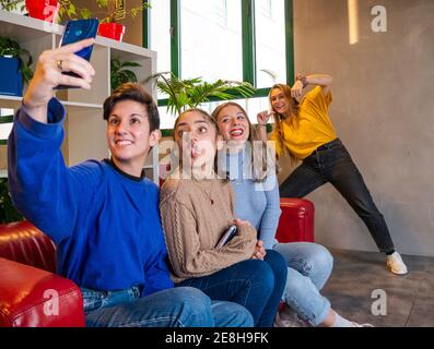 Smiling female students sitting on sofa with notebook and taking self portrait on mobile phone during break Stock Photo