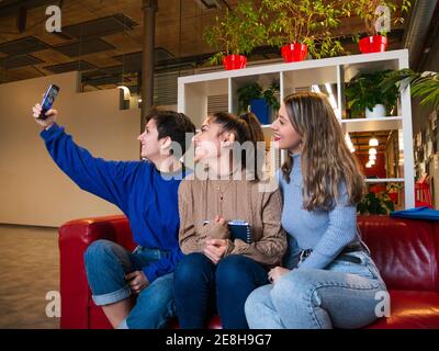 Smiling female students sitting on sofa with notebook and taking self portrait on mobile phone during break Stock Photo