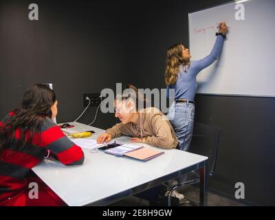 Female learner writing with marker on whiteboard while classmates working on project sitting at table in university classroom Stock Photo