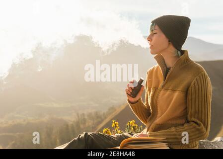 Side view of peaceful female traveler sitting on rock while smoking vape and exhaling cloud of smoke during vacation in mountains Stock Photo