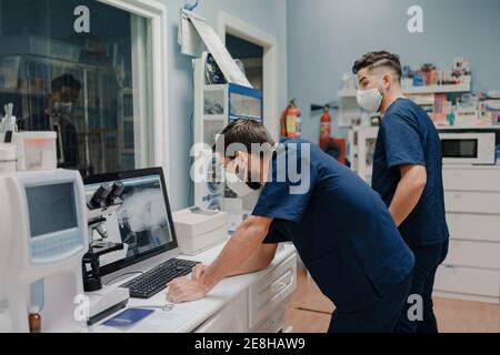 Anonymous young male doctors in respiratory masks and uniforms working on desktop computer in clinic Stock Photo