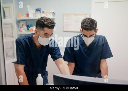Anonymous young male doctors in respiratory masks and uniforms working on desktop computer in clinic Stock Photo