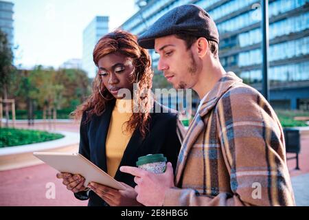 Black businesswoman with tablet speaking with male partner holding disposable glass of hot drink standing in the street