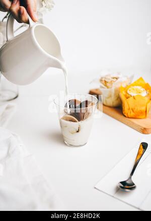 Crop anonymous person adding hot milk into glass with delicious melting chocolate bombs placed on table near sweet cupcakes Stock Photo