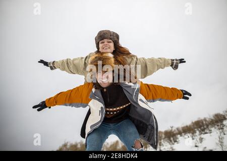 Cheerful male tourist carrying girlfriend with raised arms piggyback on snowy mount in wintertime Stock Photo