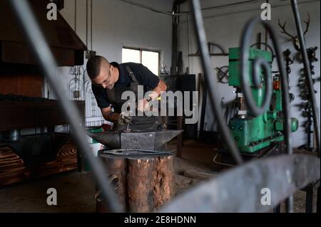 Side view of concentrated young male blacksmith in casual clothes and apron working near anvil with hammer in hand Stock Photo