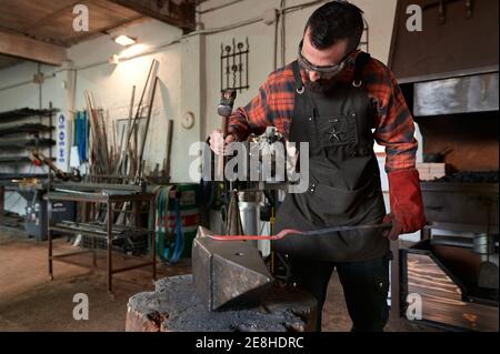Concentrated young bearded blacksmith in apron and protective goggles striking heated metal with hammer on anvil while working in forge Stock Photo