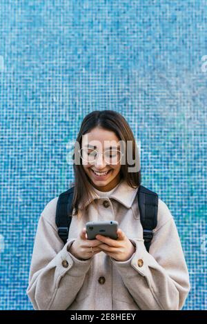 Crop attractive smiling female student in warm wear with backpack using modern mobile phone on sidewalk on street Stock Photo
