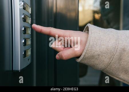 Crop anonymous female in warm coat pressing entry phone button while standing near building entrance door on clear autumn day Stock Photo