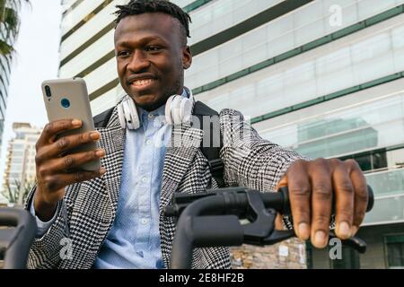 Crop glad African American male office employee with headset browsing internet on mobile phone while sitting on bike in city Stock Photo