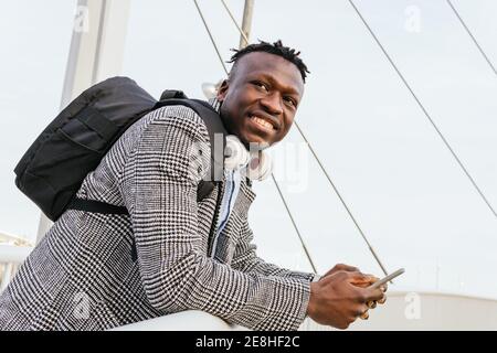 Side view of young cheerful African American male manager with backpack and cellphone leaning on fence of urban bridge while looking away Stock Photo