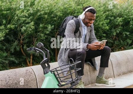 Young positive ethnic male office employee in headphones chatting on cellphone while resting near bicycle in town Stock Photo