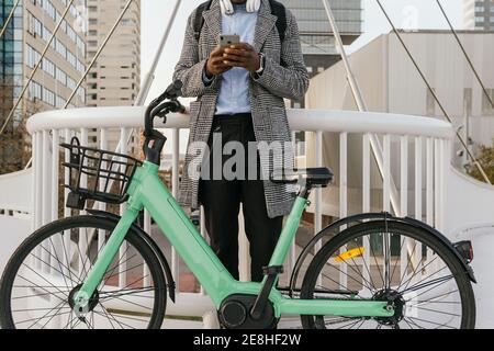 Low angle of anonymous content African American male manager in headset text messaging on cellphone near bicycle in town Stock Photo