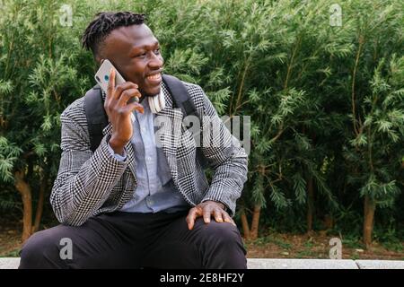 Young happy African American office worker with backpack speaking on cellphone while sitting against urban building and looking away Stock Photo