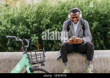 Young positive ethnic male office employee in headphones chatting on cellphone while resting near bicycle in town Stock Photo