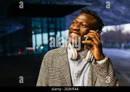 Young smiling ethnic male office worker in headphones speaking on cellphone while looking away on street in evening Stock Photo