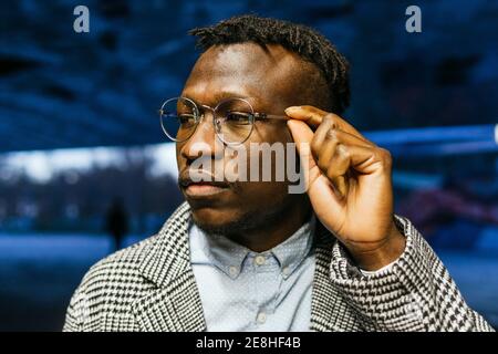 Young thoughtful African American male office employee touching eyewear while looking away in town at dusk Stock Photo