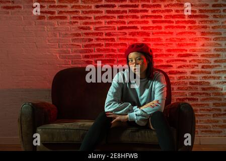 Full length confident young Asian female in stylish outfit and hat sitting on couch and looking at camera in dark room against brick wall Stock Photo