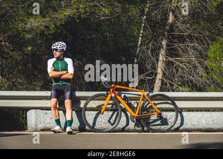Full body of young male cyclist in protective helmet and sportswear standing leaning on crash barrier near bike parked on asphalt road against blue sk Stock Photo